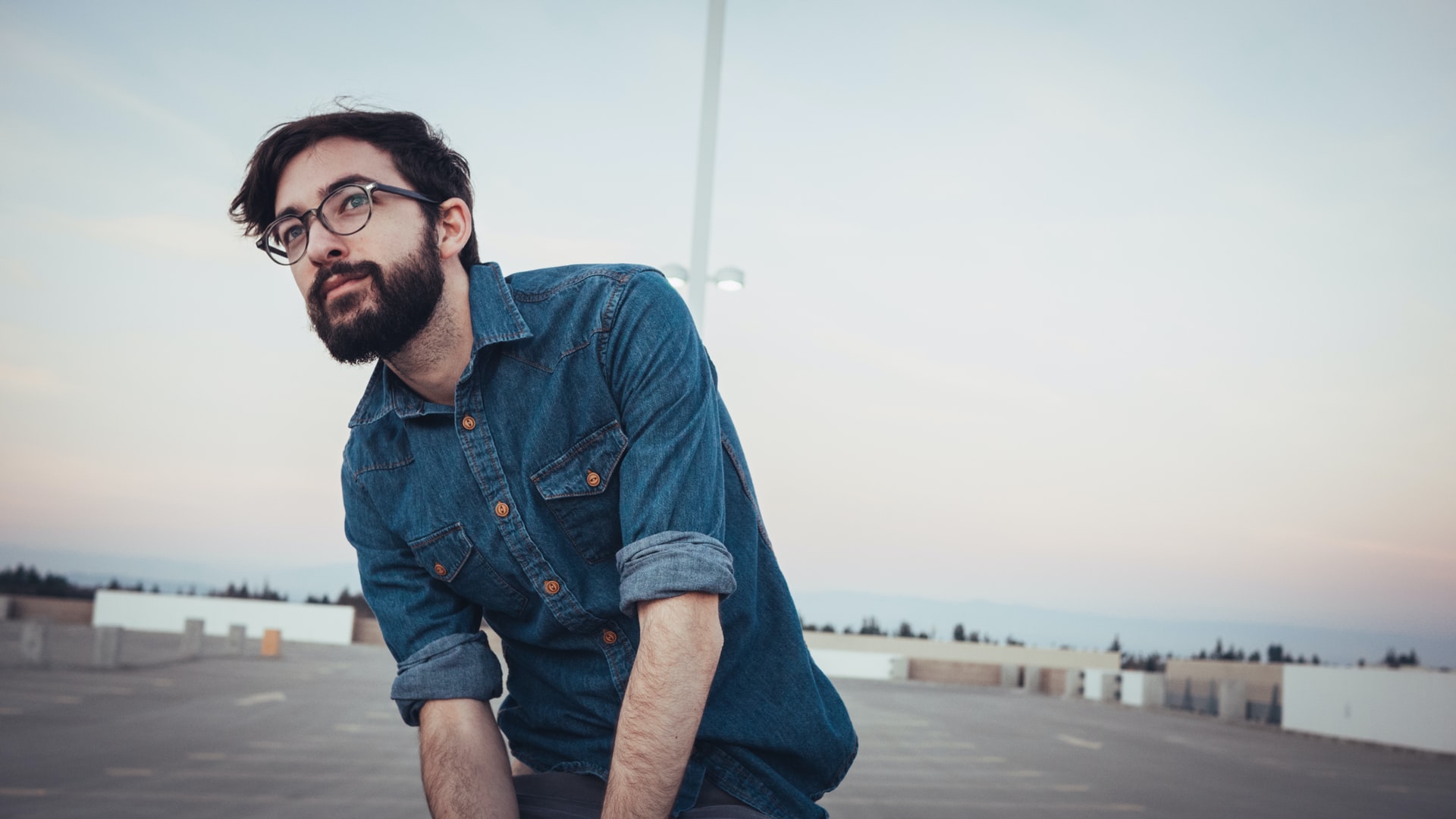 A portrait of a bearded man with jeans color shirt and eyeglasses while sitting on rooftop