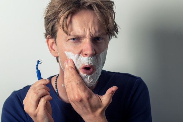A man shaving his beard with razor looking at mirror