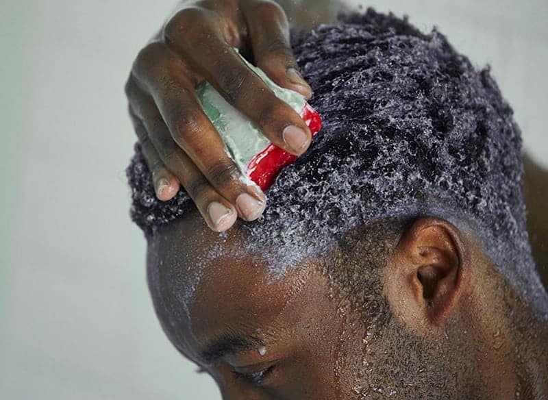 A black man applying shampoo on his curly hair while taking bath