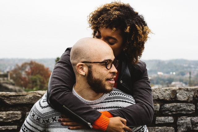 A girl hugging a man from behind and kissing the man's bald head on a outside date