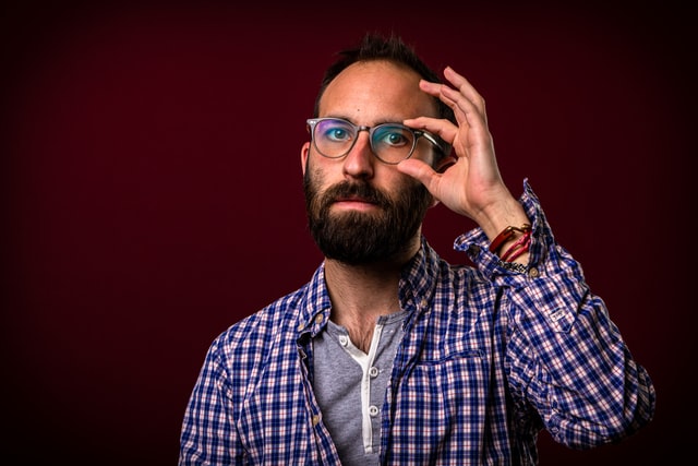 A bearded man holding his eyeglass by one hand 
